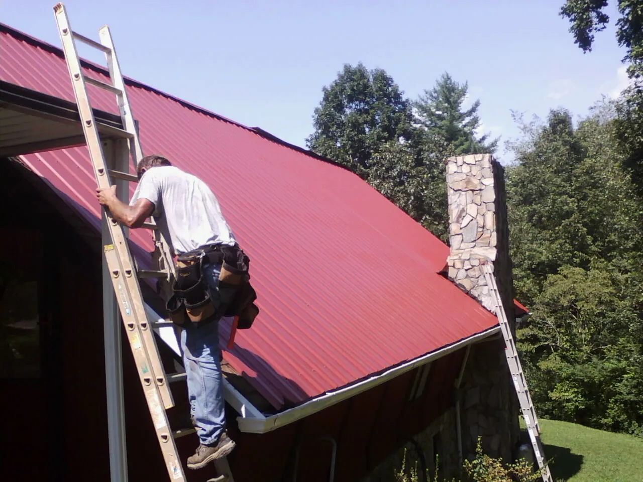 Roofer on a ladder beside a red metal roof and stone chimney — Soffit & Fascia Repair by Roofing Brothers in Walnutport